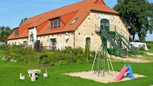 A building with a red roof, a stone facade, and a playground with slides. - 06 Apartment - Ferienhof auf Rgen (Putbus)