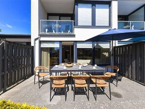 Moderni kaksikerroksinen rakennus, jossa on parveke, ruokailutila pöytineen ja tuoleineen sekä suuri aurinkovarjo.