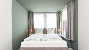 Habitación de hotel con cama, dos lámparas de noche, ventana con cortinas y vista a la ciudad.