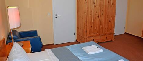 A hotel room with a bed, a wooden wardrobe, a desk with a lamp, and a blue chair.