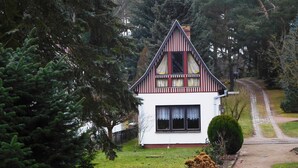 A small, single-story house with a steeply pitched roof, surrounded by dense trees and a well-maintained lawn. - Summer house, rowing boat incl. - Summer house (Krakow am See)