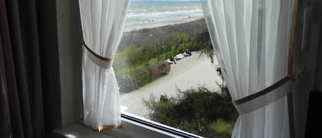 A window with sheer white curtains, a view of a beach with people walking, and a clear sky.