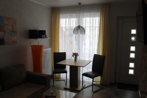 Et værelse med sofa, et bord med to stole, en radiator, et vindue med gardiner og en dør med lys.