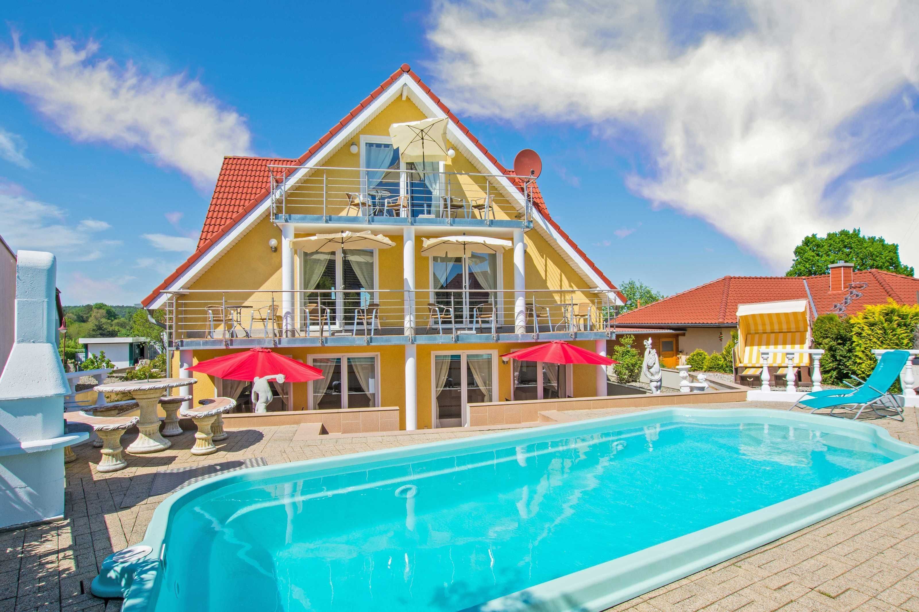 Um edifício de dois andares com piscina, área de estar ao ar livre e uma estrutura com telhado vermelho nas proximidades.