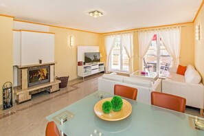 Una sala de estar con chimenea, sofás blancos, una mesa de comedor de cristal y un televisor sobre un mueble blanco.