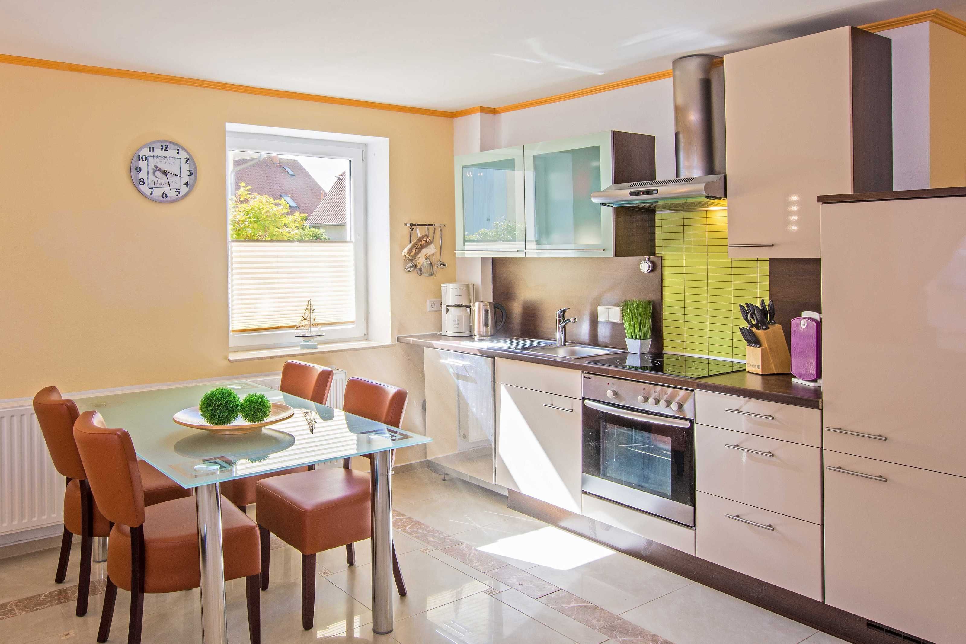 Une cuisine moderne avec un coin repas, comprenant une table en verre, des chaises marron, une horloge et une fenêtre avec des stores.