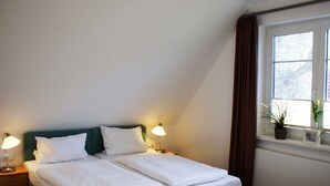 A twin bed with white linens, two bedside lamps, a green headboard, and a window with a plant on the sill. - Double room 08 - Guest house & beach hall (Ahrenshoop)