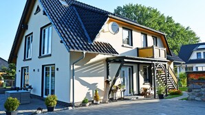 Een huis met twee verdiepingen, een schuin dak, een balkon en een kleine tuin met potplanten.