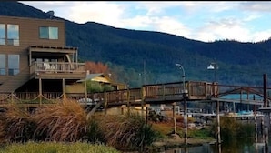 Un edificio moderno su due piani con un balcone, un pontile in legno e un ponte sopra una distesa d'acqua, circondato da montagne.