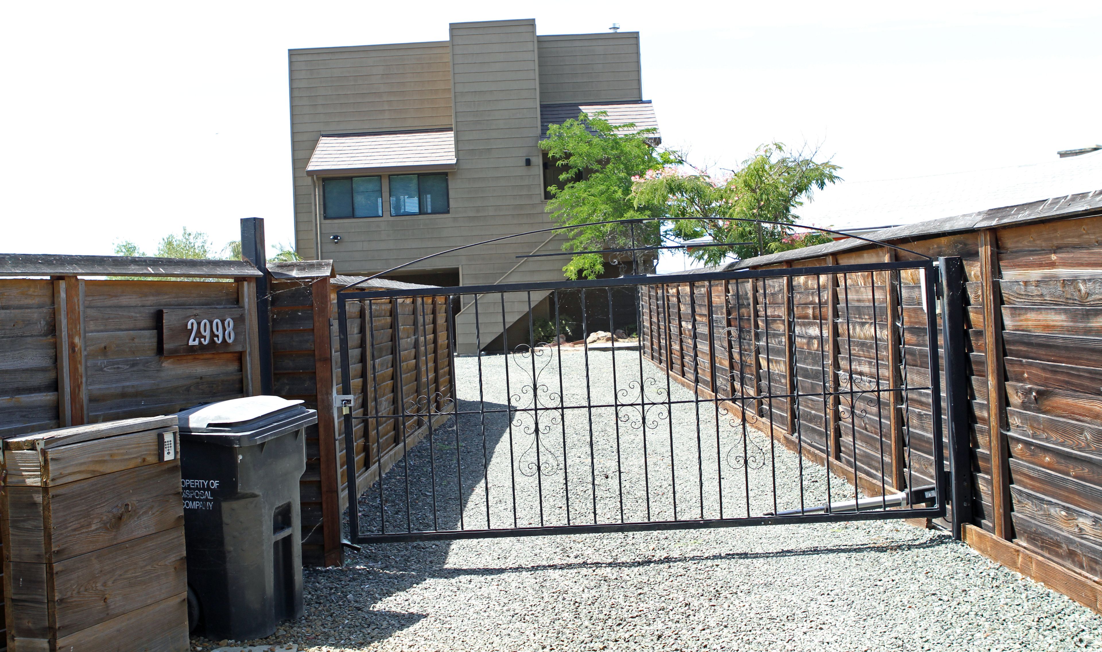 Une entrée fermée par un portail, donnant sur un immeuble à l’architecture moderne, une clôture en bois et une allée gravillonnée.