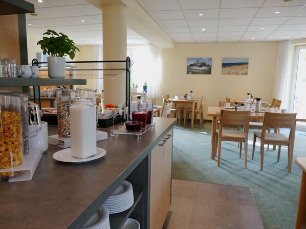 A breakfast bar with cereal, milk, and juice options, surrounded by wooden tables and chairs in a bright room with large windows.