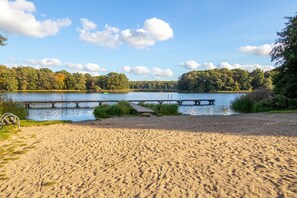 Een zandstrand met een steiger die uitsteekt in een kalm meer, omringd door dichte bomen.