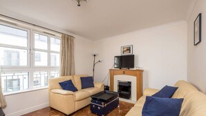 A living room with two beige sofas, a small table, a fireplace, a television, and a window with curtains. - Old Town Flat with Parking near Royal Mile (Edinburgh)