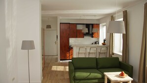 Ein modernes Wohnzimmer mit einem grünen Sofa, einem kleinen Tisch mit Obst und einer Küche im Hintergrund.