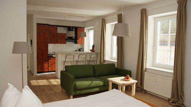 Ein Hotelzimmer mit einem grünen Sofa, einem kleinen Tisch mit Früchten, einem Bett, einer Küchenzeile und einem Fenster mit Vorhängen.