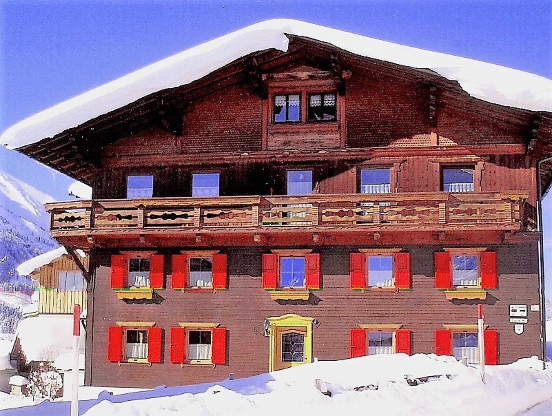 A multi-story wooden building with red shutters, a balcony, and a mountain backdrop.