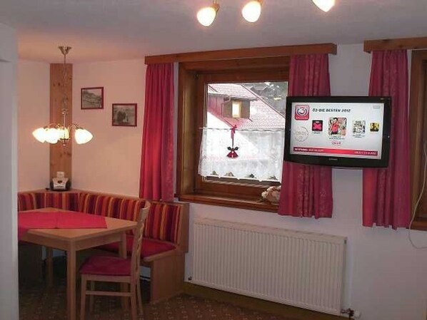 Quarto com mesa de jantar, cadeiras, janela com cortinas, TV na parede e radiador.
