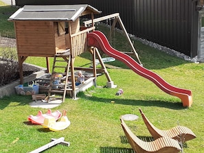 A wooden playhouse with a slide, a small deck, and a garden area with toys and a toy car.