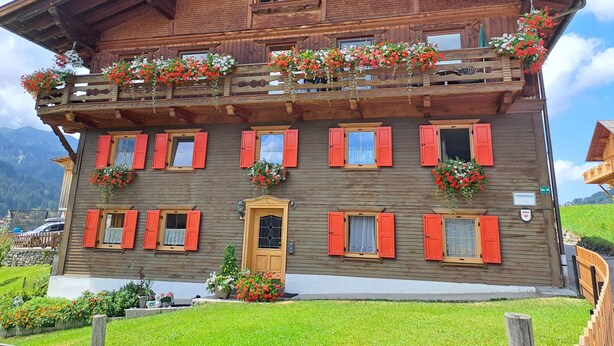 Un edificio de madera de varios pisos con balcones, contraventanas rojas y macetas con flores.