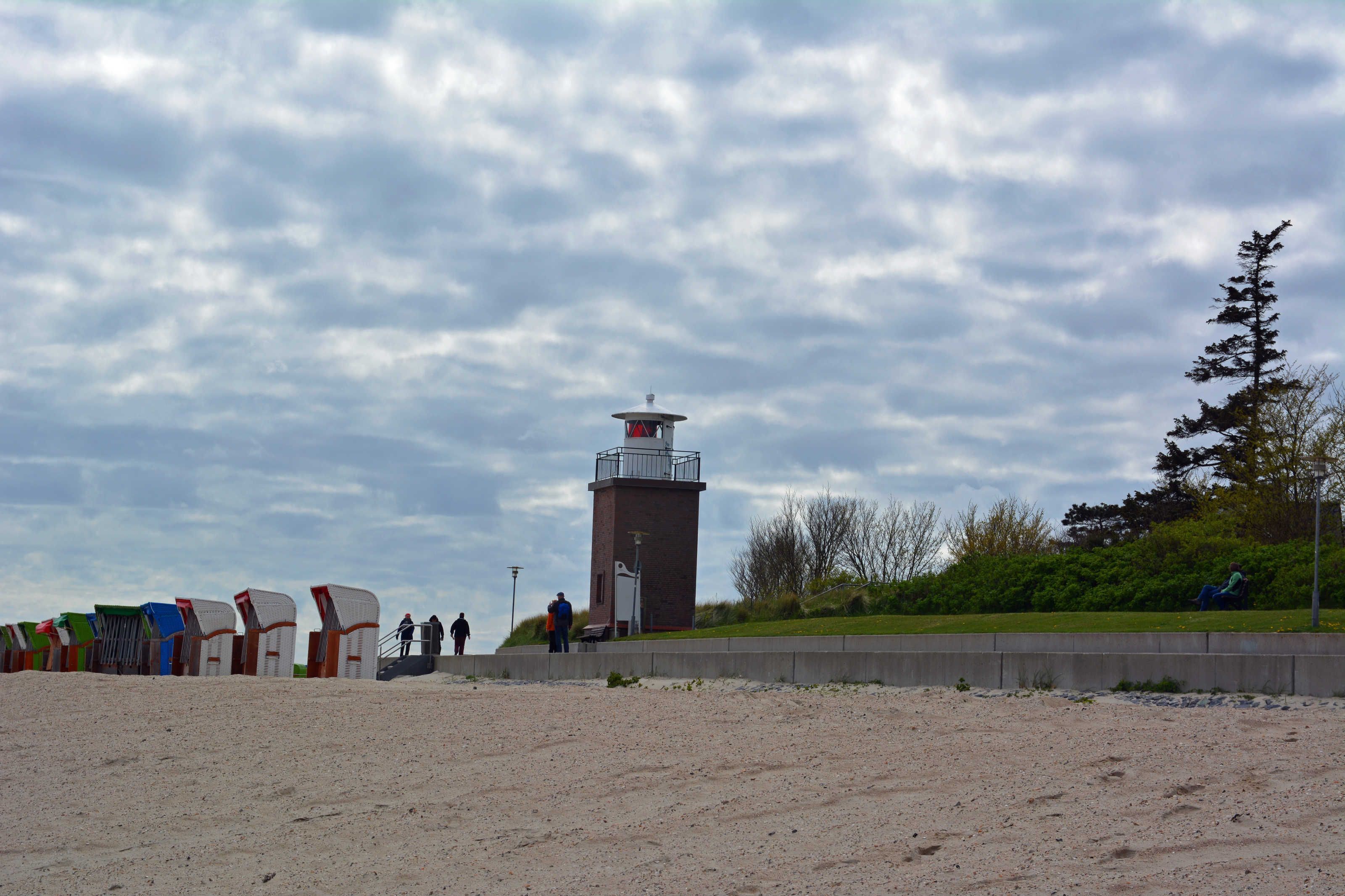 Uma praia com um farol, uma fileira de cabanas de praia e uma área gramada com árvores.