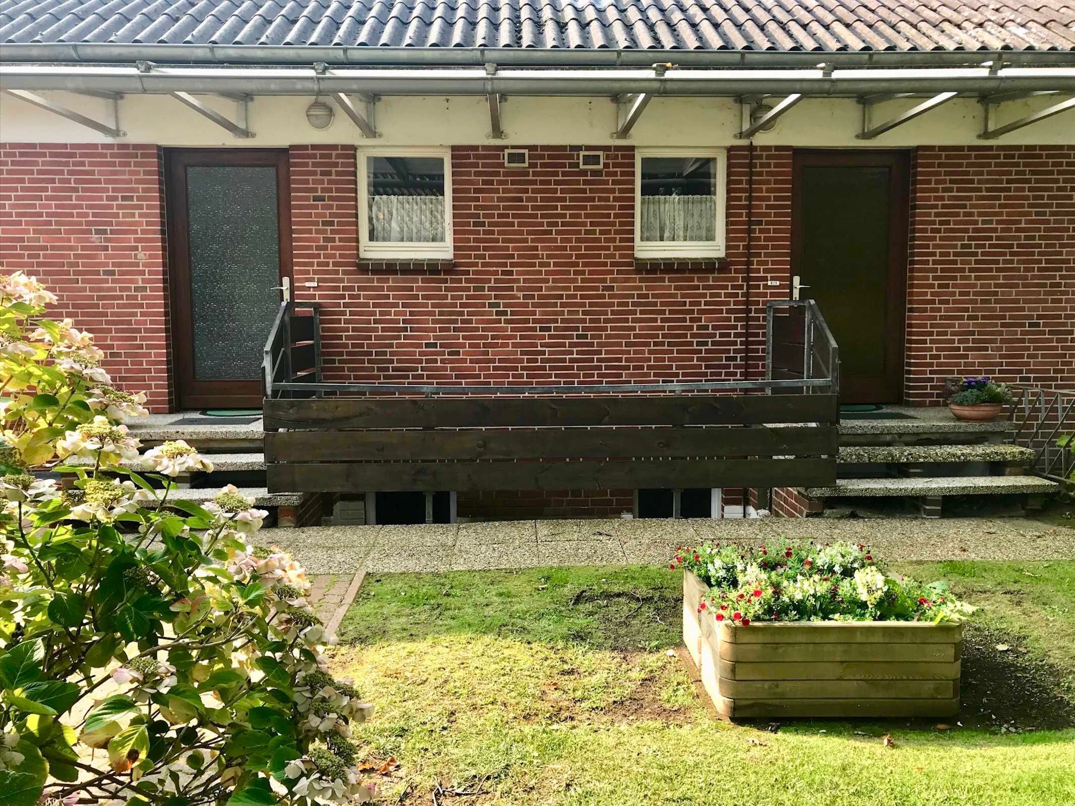 Une maison en briques avec un porche à l’avant, deux fenêtres aux cadres blancs et un petit jardin agrémenté d’un bac à fleurs en bois.
