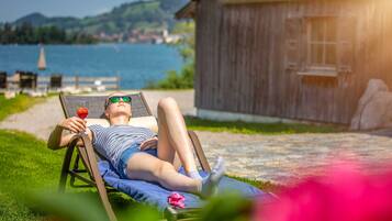 A person relaxing on a lounge chair with a drink, overlooking a lake and a wooden building.