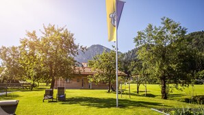 A flagpole with a flag, a building with a red-tiled roof, and a grassy area with trees and a mountain in the background.