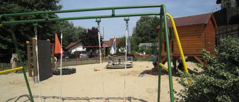 Ein Spielplatz mit Schaukeln und einer Rutsche, umgeben von Sand und viel Grün.