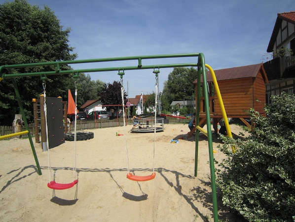 Ein Spielplatz mit Schaukeln und einer Rutsche, umgeben von Sand und viel Grün.