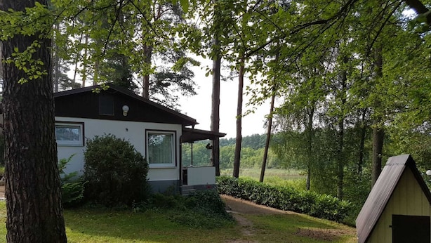 Ein Haus mit weißer Fassade, einer Laube und einem kleinen Gartenhaus in einer bewaldeten Gegend.