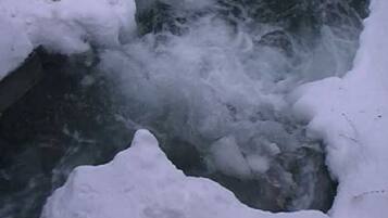 Schnee liegt in der offenen Landschaft, ein Bach fließt teilweise gefroren hindurch.