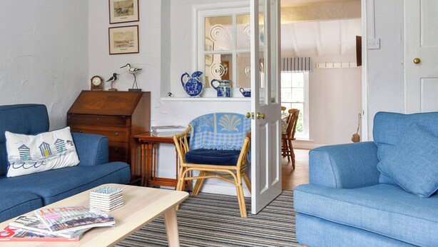 Ein Wohnzimmer mit zwei blauen Sofas, einem Couchtisch mit Zeitschriften, einem Korbstuhl und einer Tür zu einem Küchenbereich mit einem Fenster.