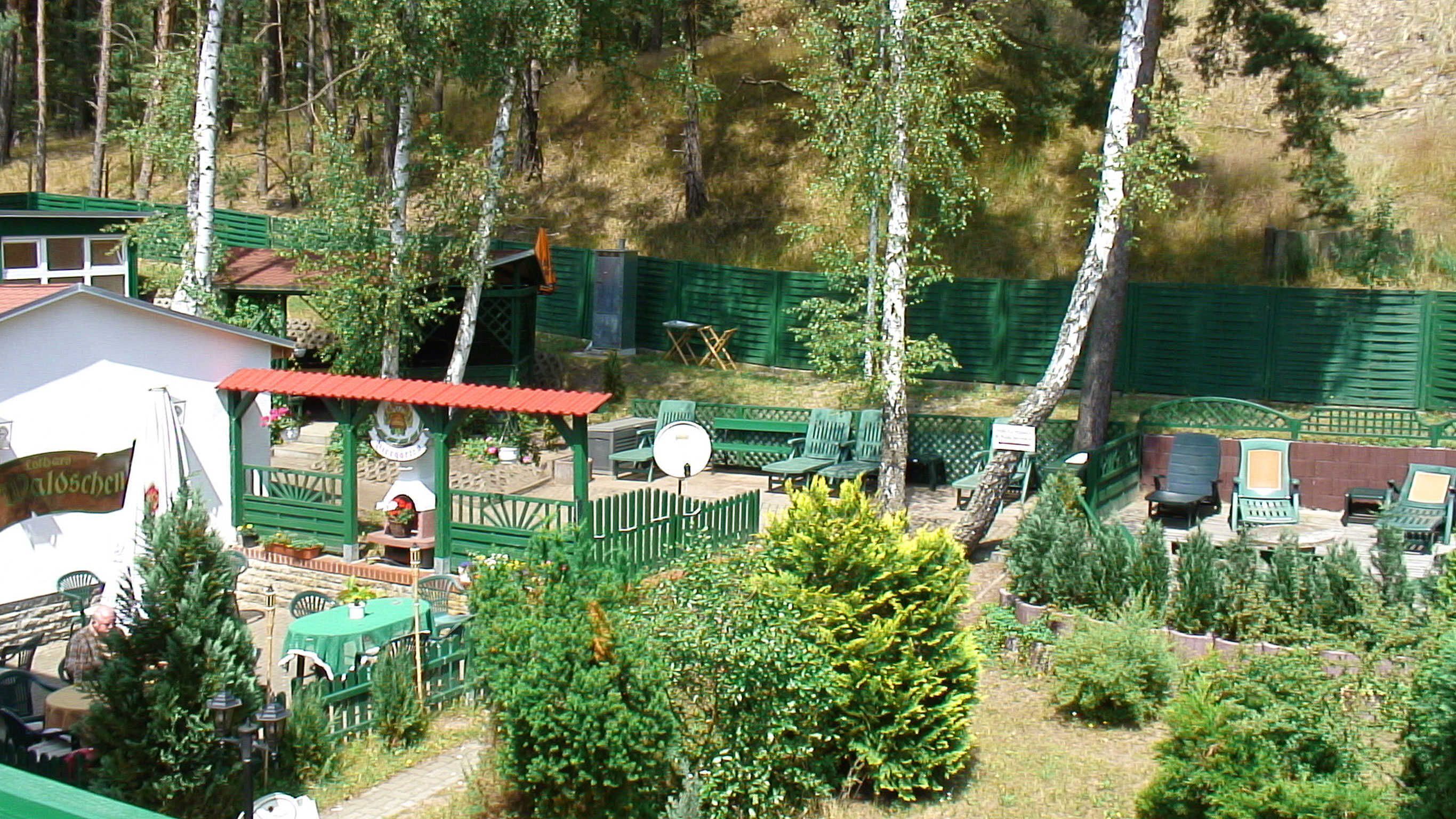 A rustic outdoor seating area with green furniture, surrounded by trees and a small building with a red roof.