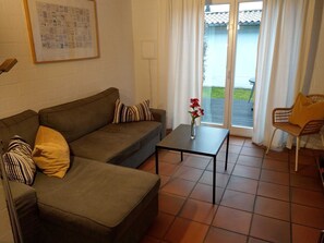 Una sala de estar con un sofá, una mesa de centro y una silla. Se ve una ventana con cortinas, una puerta y un piso con baldosas.