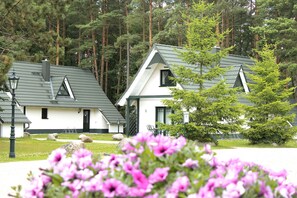 A row of white cabins with dark roofs, surrounded by a forest. - 95m² holiday home - holiday homes on the lake (Nossentiner Hütte)