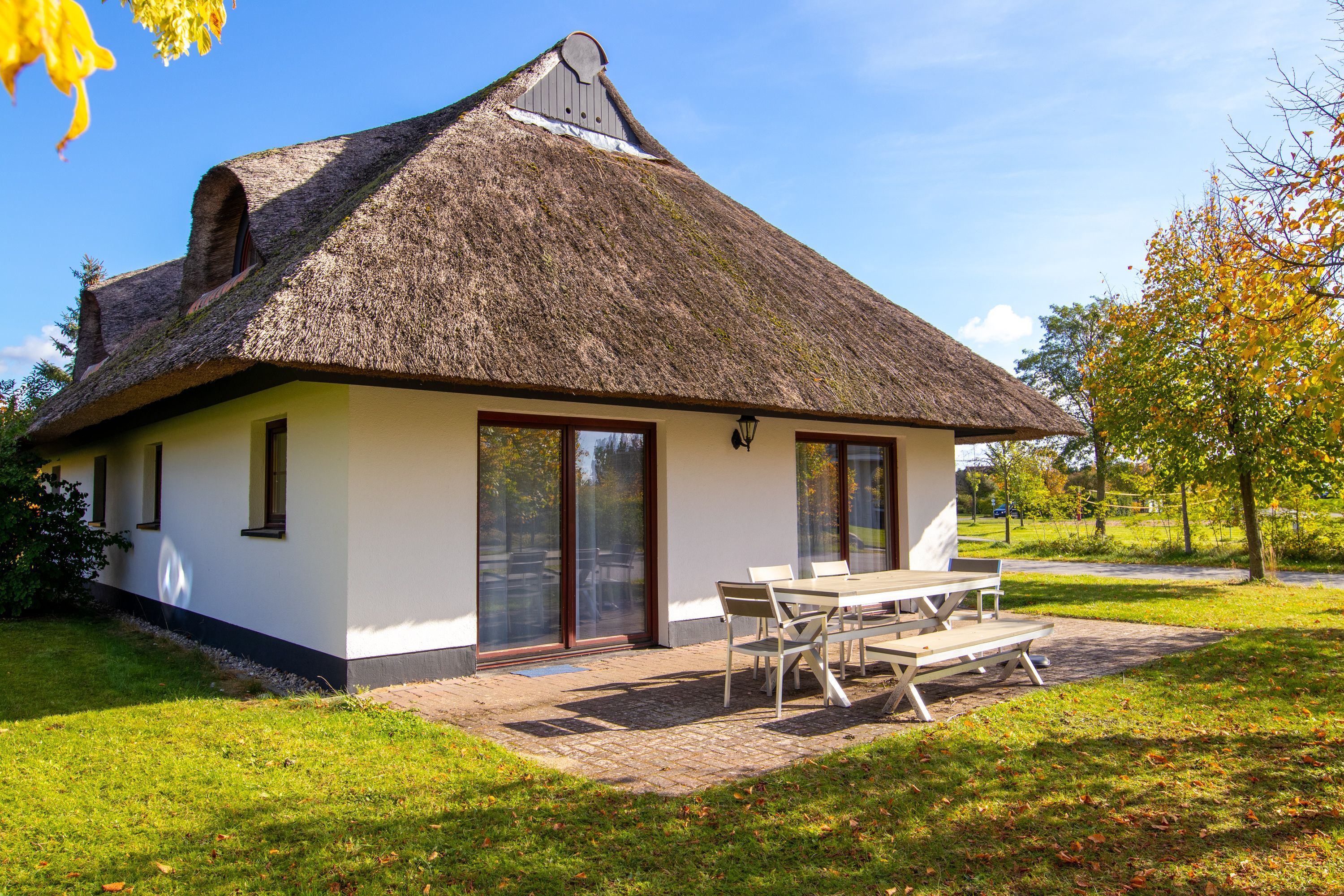 Ein Haus mit Strohdach, großen Fenstern und einer Terrasse mit Tisch und Stühlen.