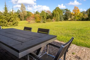 Una mesa de comedor con sillas, sobre una zona empavedada con un campo de césped y árboles al fondo.