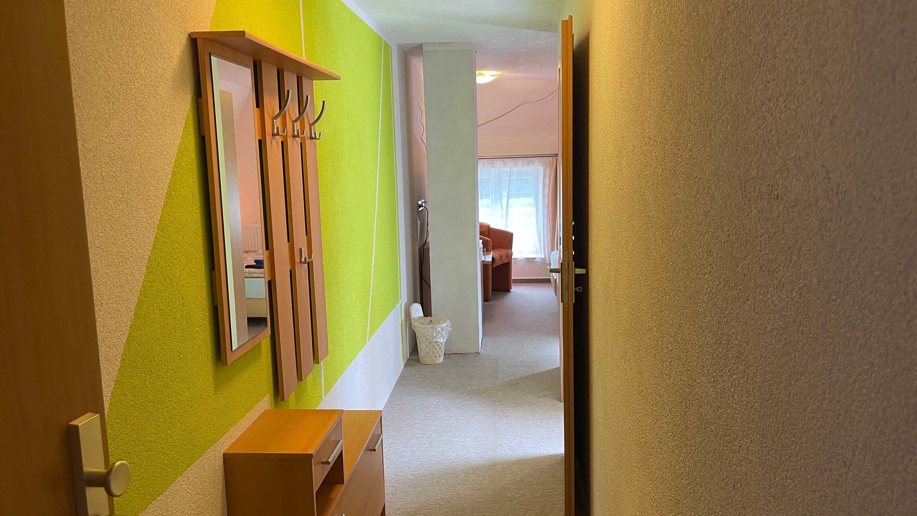 A hallway with a mirror, a wooden cabinet, and a door leading to a room with a sofa and a window.