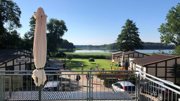 A view from a balcony overlooking a lake, with a building labeled 'Ferlenhäuser' and a parking area with cars. - 08 DZ - Island-Hotel Dobbertin (Dobbertin)