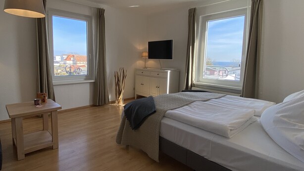 Habitación de hotel con cama, mesita de noche, lámpara, televisor y ventanas amplias con cortinas.