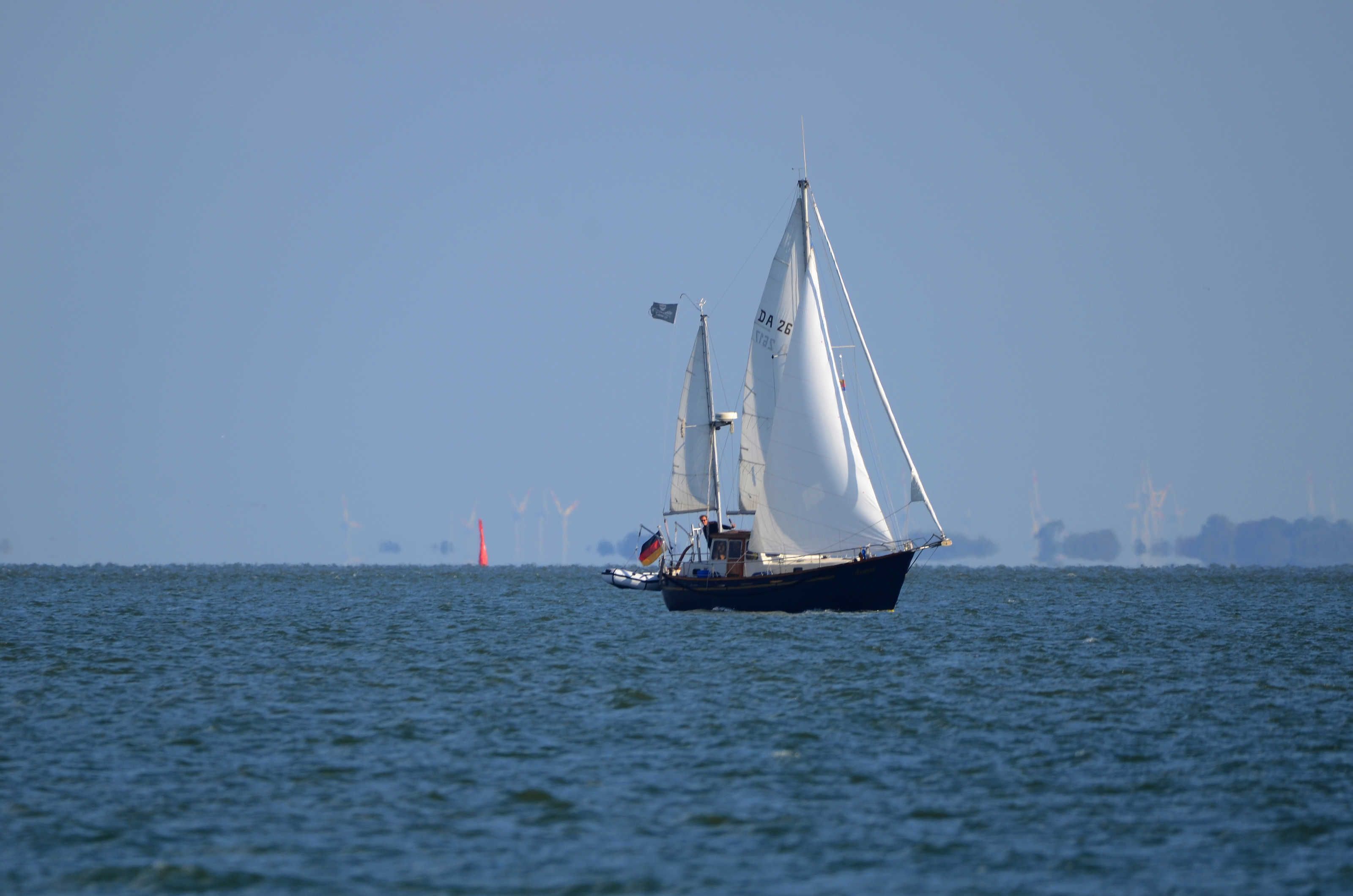 Un voilier sur une mer calme, avec une côte lointaine et des éoliennes en arrière-plan.