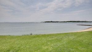 Un paisaje costero con un primer plano de hierba, una playa arenosa y un mar tranquilo con barcos.