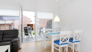 A modern living room with a black sofa, white dining table with blue cushions, and a large window overlooking a brick building. - Vacation apartment Leuchtturm - Haus Kunterbunt (Esens)