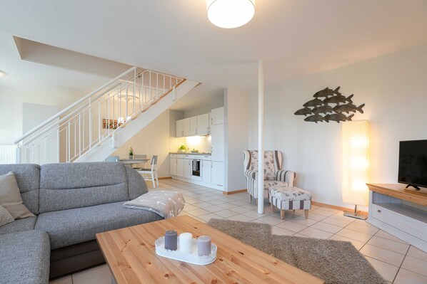 A modern living room with a grey sofa, a wooden coffee table with candles, a staircase, a kitchen in the background, and a wall-mounted fish decoration.