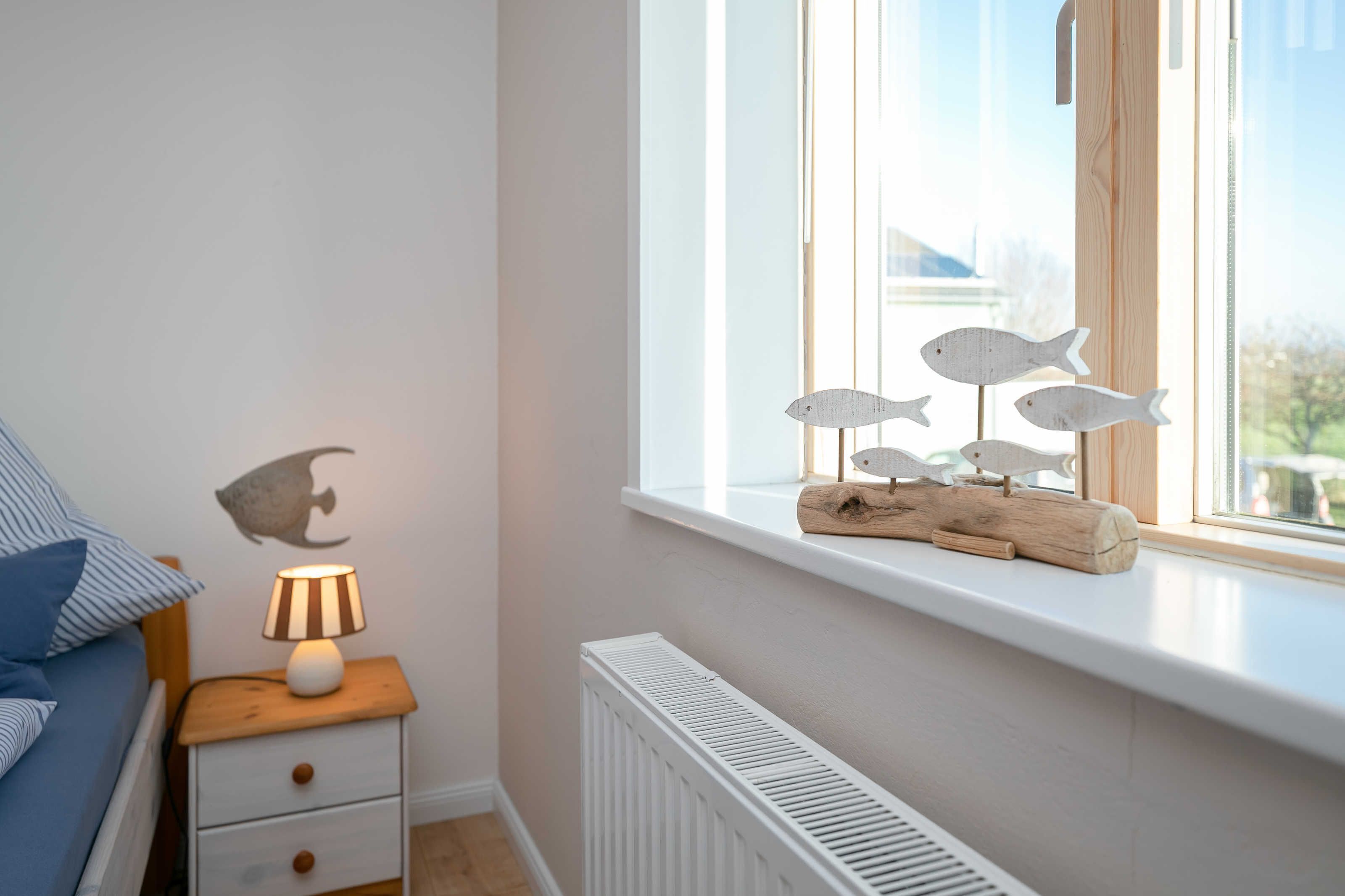 A bedroom with a bed, a nightstand with a lamp, a radiator, and a window with a wooden shelf holding fish-shaped decorations.