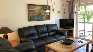 A living room with a black leather sofa, a wooden coffee table, a painting of a seascape, a TV, and a window with curtains.
