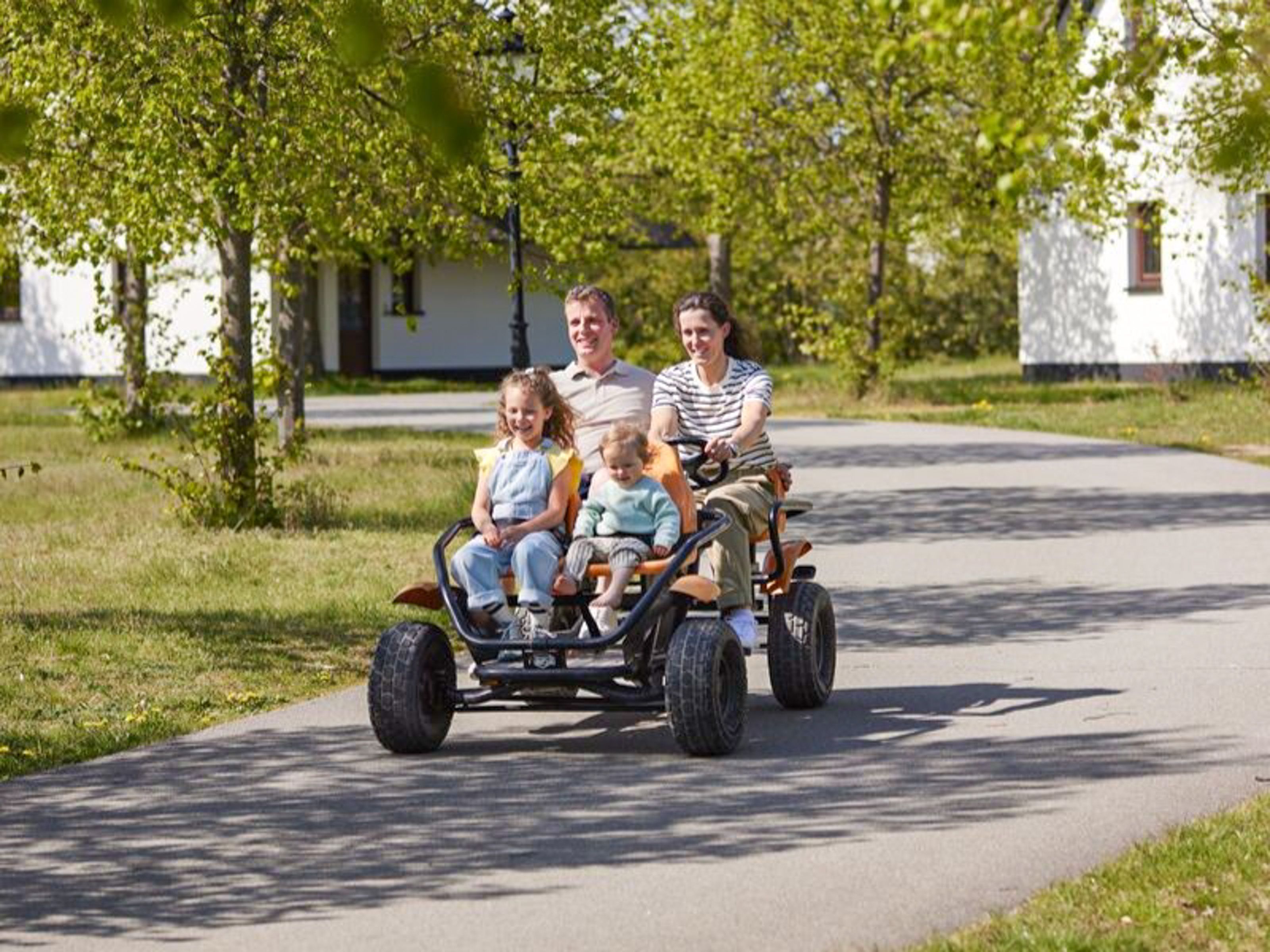 A family on a three-wheeled vehicle on a paved path with trees and houses in the background.