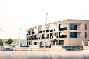 A modern multi-story building with balconies, outdoor seating, and a waterfront view. - The Edged (06) - Z1-THE HOUSE (Zwenkau)
