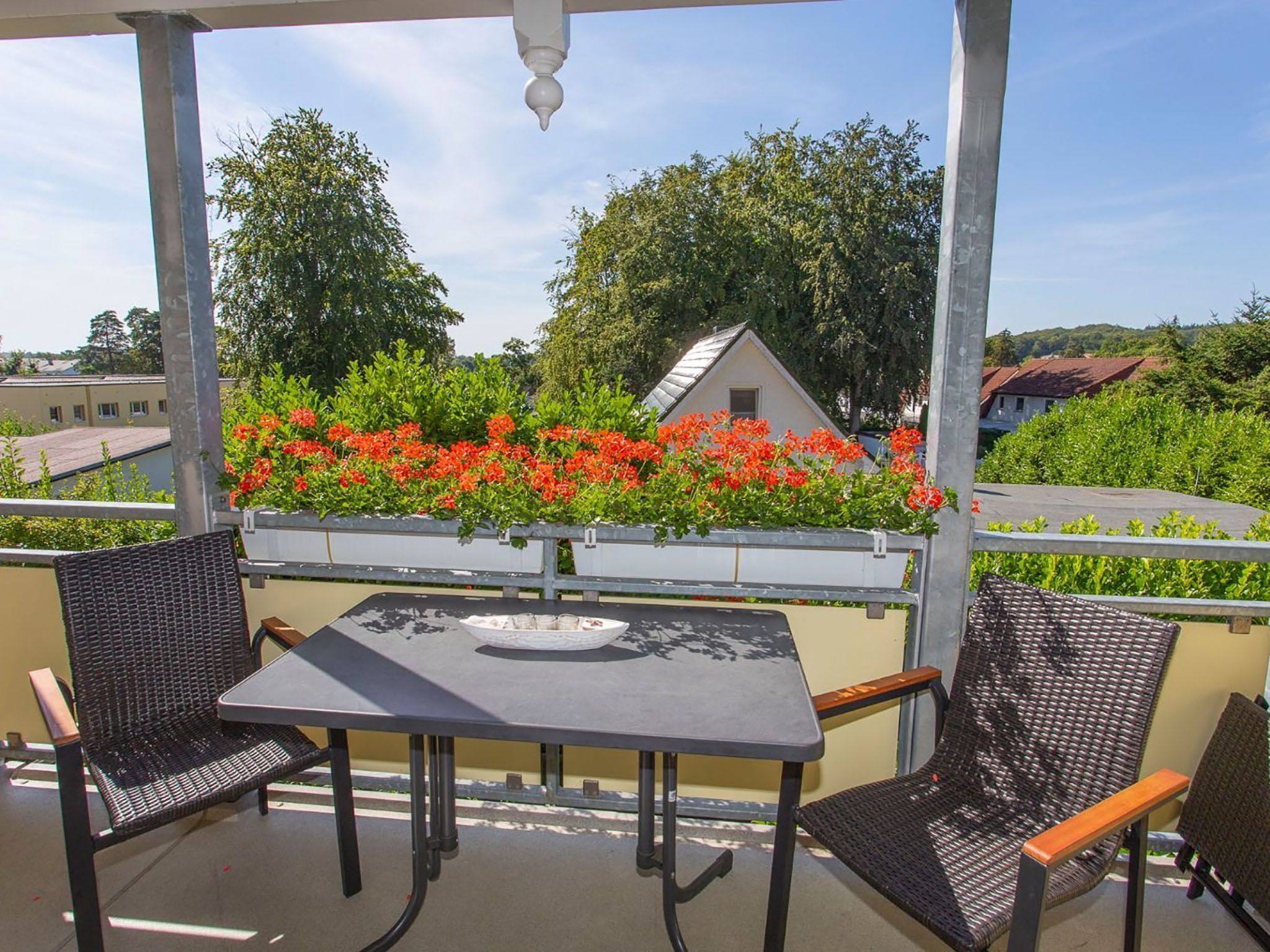 Un espacio de estar al aire libre cubierto con dos sillas y una mesa, con vistas a un jardín con flores rojas y un edificio al fondo.