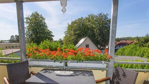 Ein überdachter Außenbereich mit zwei Stühlen und einem Tisch, mit Blick auf einen Garten mit roten Blumen und einem Gebäude im Hintergrund.